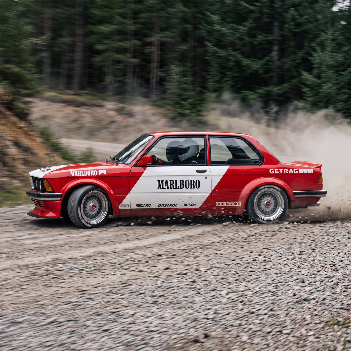 Un BMW Serie 3 E21 rojo y blanco de rally deslizando sobre una pista de grava en un bosque, levantando polvo y piedras, con dos ocupantes con casco