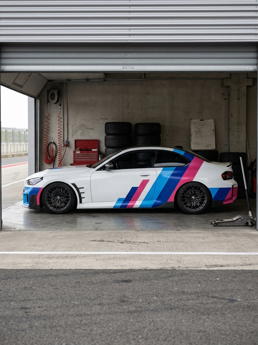 Un BMW blanco con librea de competicion en azul y magenta, parado dentro del box de un circuito con neumaticos apilados al fondo y el pit lane delante
